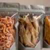 三只自立拉链袋装着虾干、鱿鱼丝和鱼排零食，展示水产干货包装袋的防潮防腥选型场景