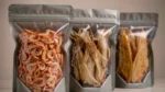 三只自立拉链袋装着虾干、鱿鱼丝和鱼排零食，展示水产干货包装袋的防潮防腥选型场景