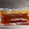 不锈钢台面上放着一个表面泛着红油光泽的柔性零食包装袋,周围散落着辣椒碎和芝麻